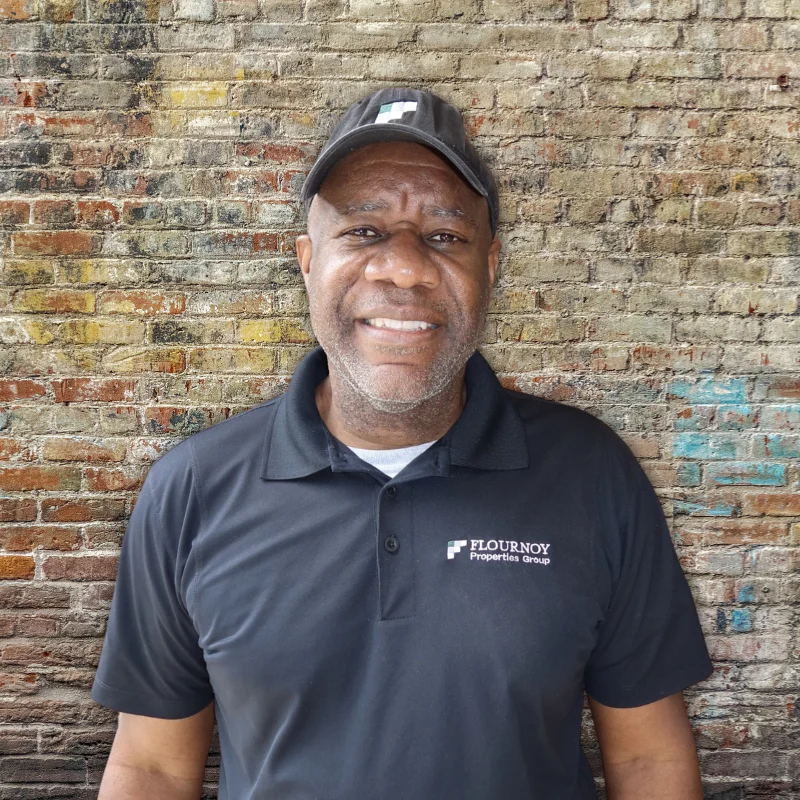 A man wearing a black polo shirt and cap stands in front of a rustic brick wall, smiling confidently.