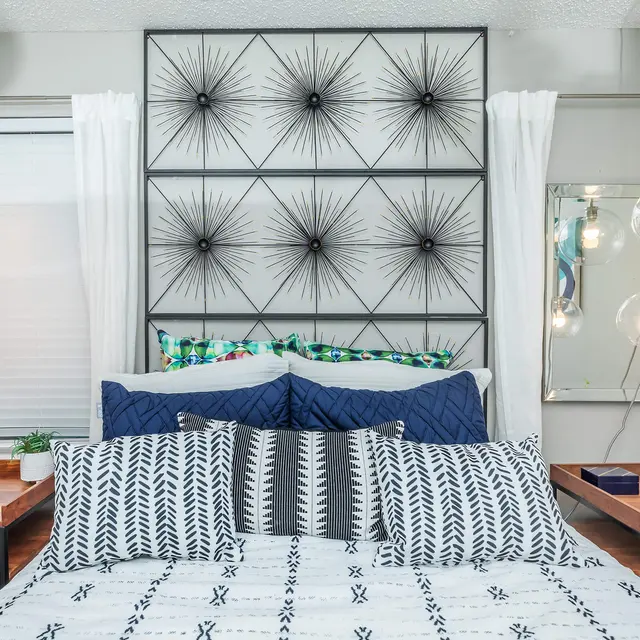 A modern bedroom featuring a stylish headboard with a geometric design, two nightstands with decor, and bright pendant lighting, complemented by light curtains and decorative pillows on the bed.