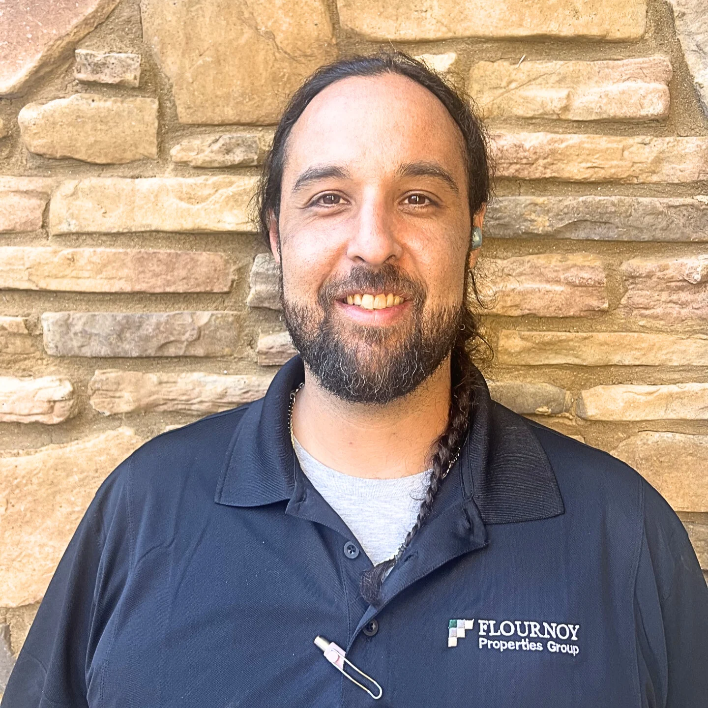 Smiling man in front of stone wall A man with long hair and a beard smiling, wearing a black shirt with a logo on it, standing in front of a textured stone wall.