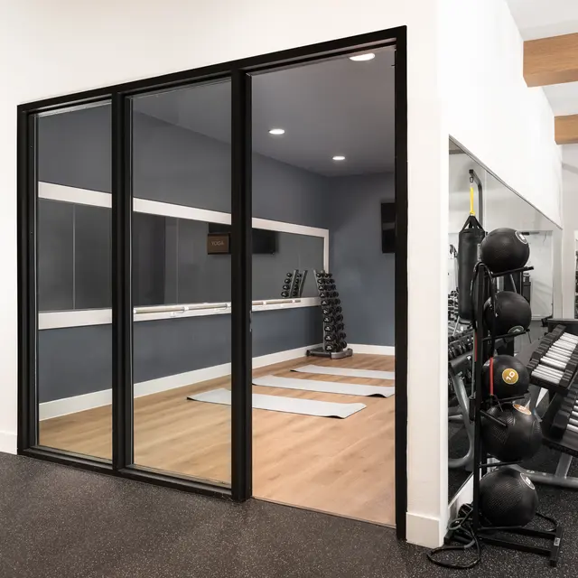 A view of a modern gym with a workout area separated by glass walls. The space features exercise mats on a wooden floor, mirrored wall sections, and various gym equipment including free weights and a punching bag.