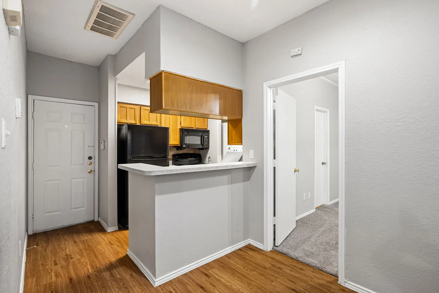 Compact Kitchen Design View of a small kitchen area with a bar countertop, featuring wooden cabinets, a black refrigerator, and a microwave. The entrance door is visible, along with a door leading to another room.