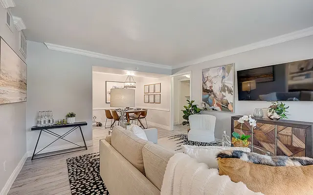 A modern living room featuring a neutral color palette, comfortable seating, and stylish decor. The layout includes a soft sofa with throw blankets, a coffee table with decorative items, and a flat-screen TV on the wall. To the side, there is a dining area with elegant chairs and a pendant light fixture.
