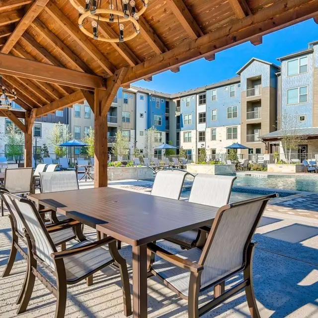 A spacious outdoor area with a wooden pergola featuring a long dining table and chairs, adjacent to a swimming pool and modern apartment buildings.