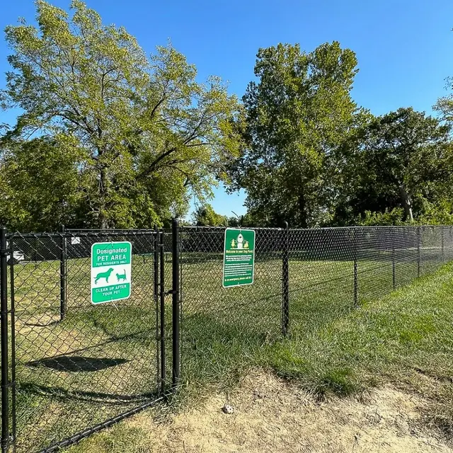Dog Park Area A fenced dog park area with signs indicating it's a designated pet area. The park features green grass and trees in the background under a clear blue sky.