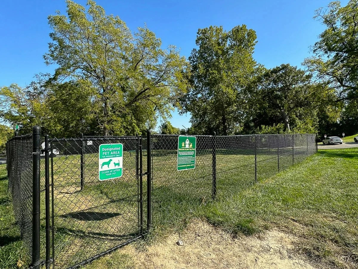 A fenced dog park area with signs indicating it's a designated pet area. The park features green grass and trees in the background under a clear blue sky.