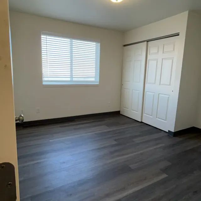 A clean, empty room with gray laminate flooring and white walls. A window with horizontal blinds allows natural light in, and there are two closed closet doors on one wall. A ceiling light fixture provides additional lighting.