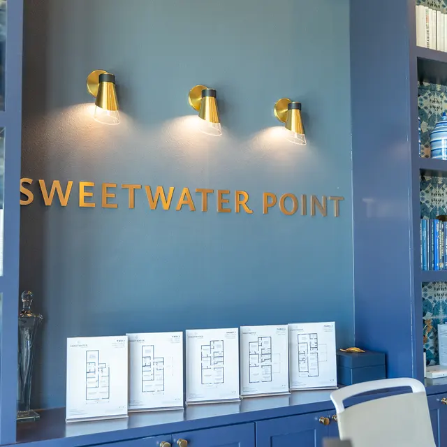 A decorative interior wall featuring the name 'Sweetwater Point' in gold letters. Below, there are architectural blueprints displayed on a shelf, along with blue and white ceramics and assorted decor. The background has a patterned blue wallpaper and wall-mounted lights.