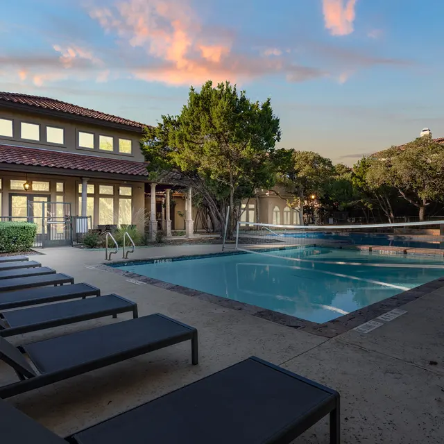Outdoor swimming pool area with lounge chairs and a clubhouse in the background, surrounded by trees and a beautiful sky