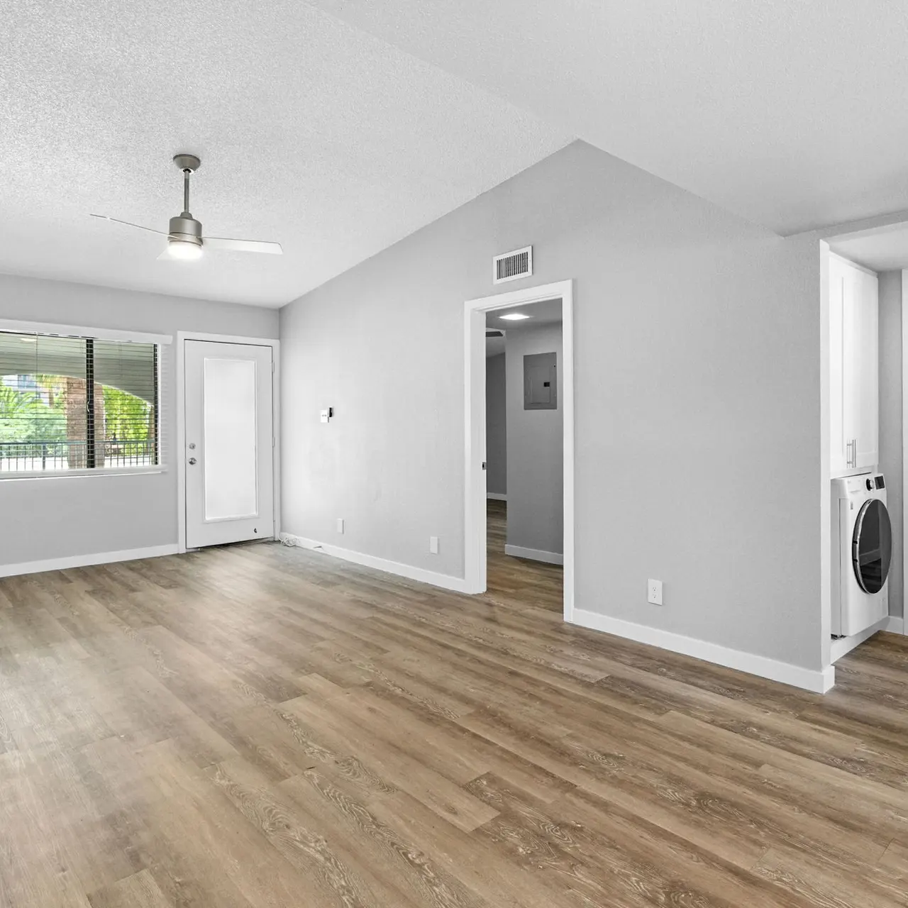 Modern Living Room Design A spacious and modern living room with light gray walls and wooden flooring. There is a ceiling fan and large windows allowing natural light. A door leads outside to a balcony. On the right, there's a laundry area with a washer and dryer.