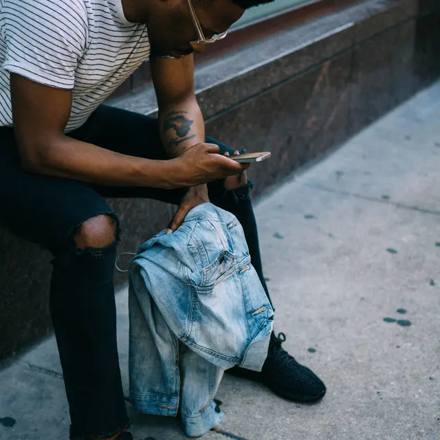 A man sitting on the ground, looking at his phone while holding a denim jacket. He is wearing a striped t-shirt and black jeans, with sunglasses on.