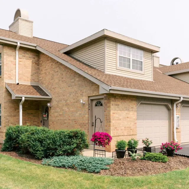A brick townhouse with a garage, surrounded by green grass and colorful flower beds. The building features a sloped roof and multiple windows.