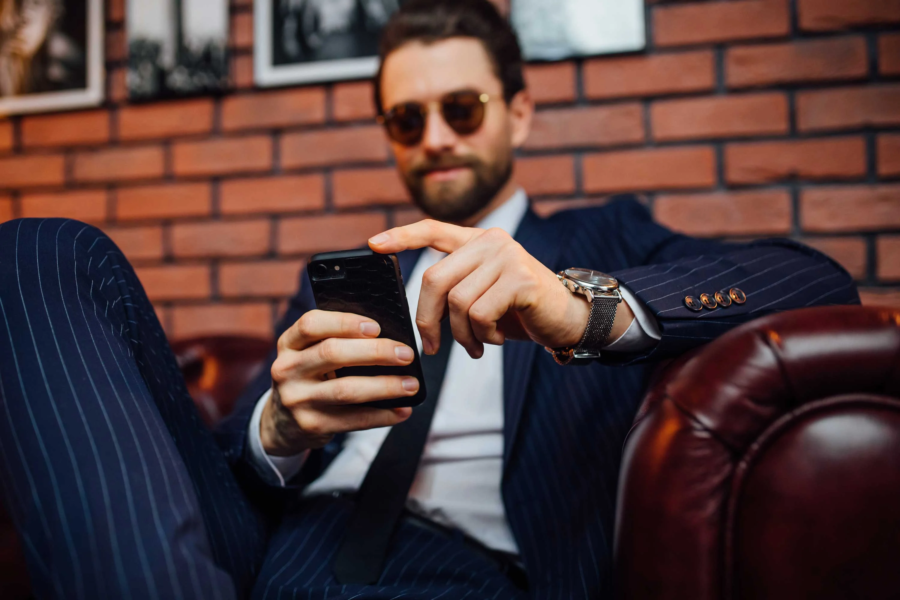 Businessman Relaxing with Smartphone A man in a striped suit and sunglasses sitting on a leather couch, looking at his smartphone.