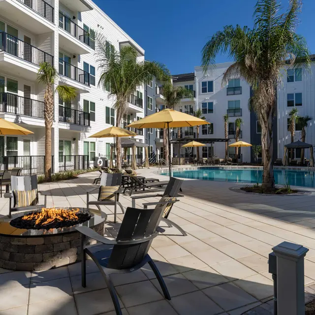 A modern apartment complex with a pool area featuring lounge chairs and umbrellas. A fire pit is situated in the foreground.