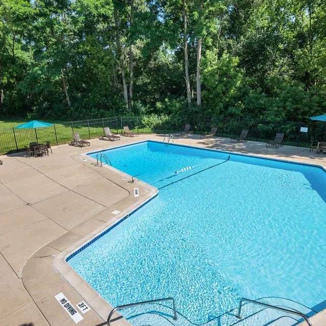 A clear blue swimming pool surrounded by a concrete patio and lush greenery. Several lounge chairs are placed around the pool, and a few teal umbrellas provide shade.