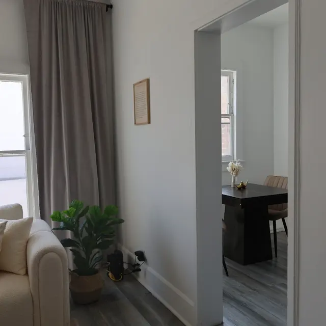 A bright living room with a light-colored couch and large windows. The couch is adorned with cushions. To the side, a potted plant adds greenery. A doorway leads to another room with a dining table and chairs visible in the background.