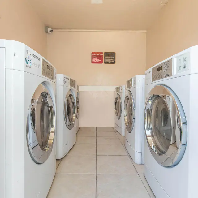 A clean and bright laundry room featuring six white washing machines arranged in a row, along with tiled flooring and light-colored walls.