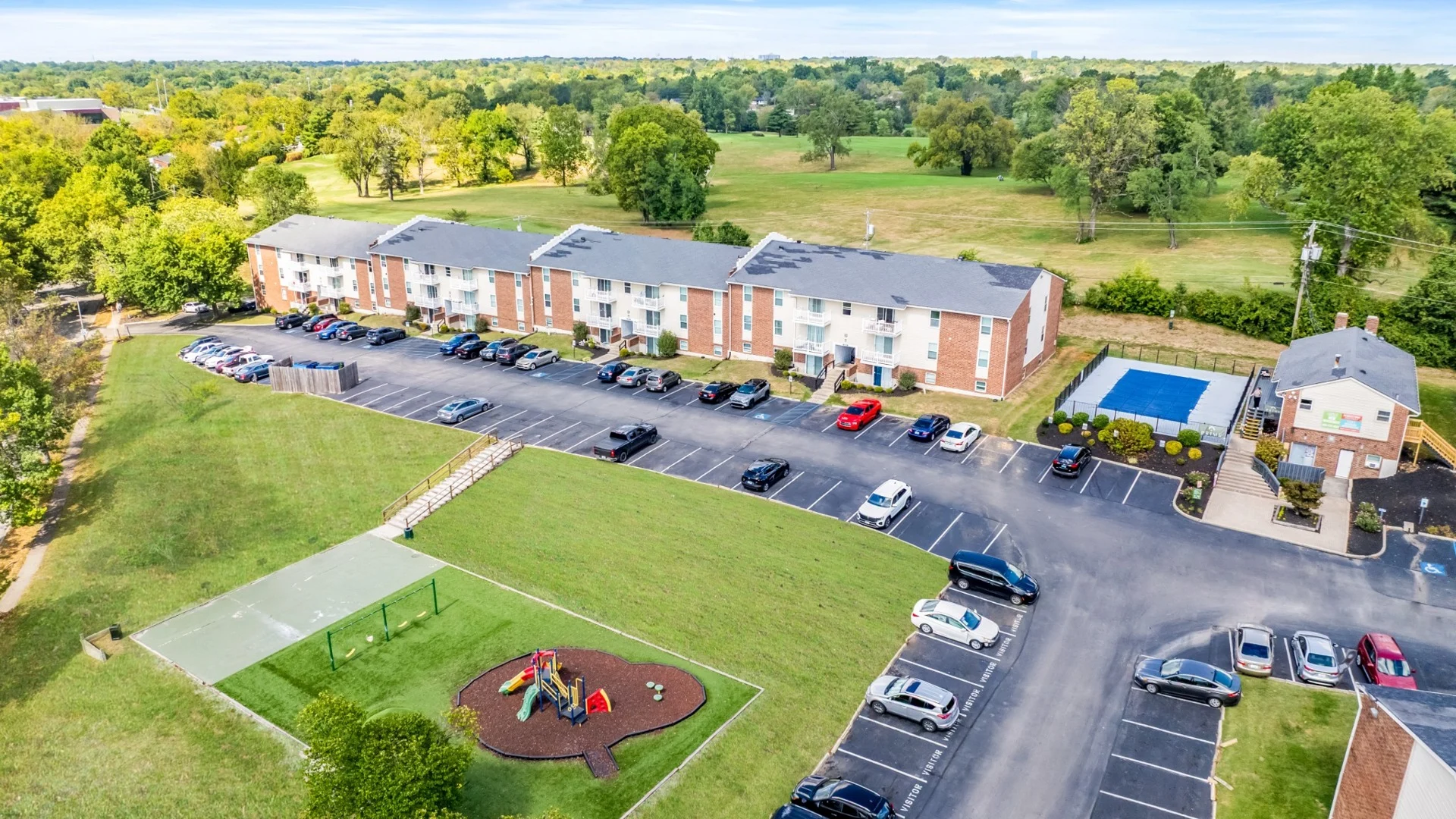 Prime at Lexington Apartment Homes aerial view Aerial view of an apartment complex with parking, playground, and pool, surrounded by green trees and grassy areas.