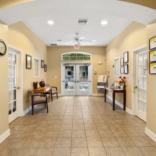 Interior hallway of a lobby with tile floor, with doors at the end leading outside, framed pictures on the walls, and furniture on either side.