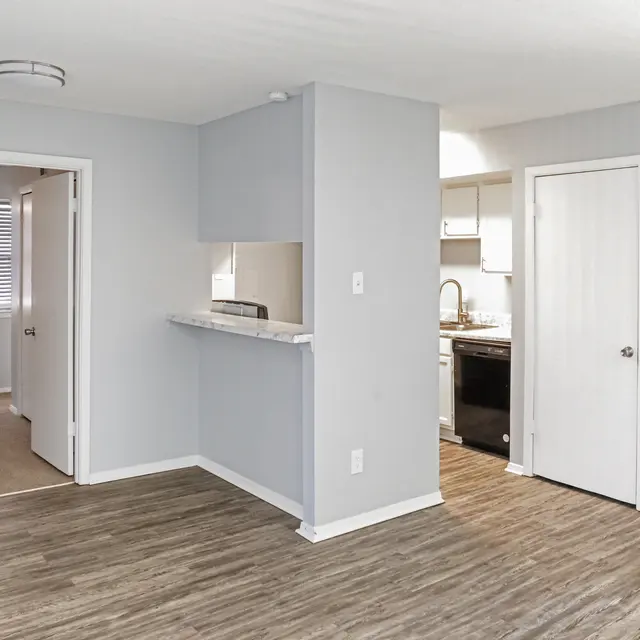 A modern apartment interior showcasing an open floor plan with light gray walls, hardwood-style flooring, and ambient lighting. An open kitchen is visible to the right with white cabinets. Two doors lead to additional rooms, and a small counter area separates the kitchen from the living space.