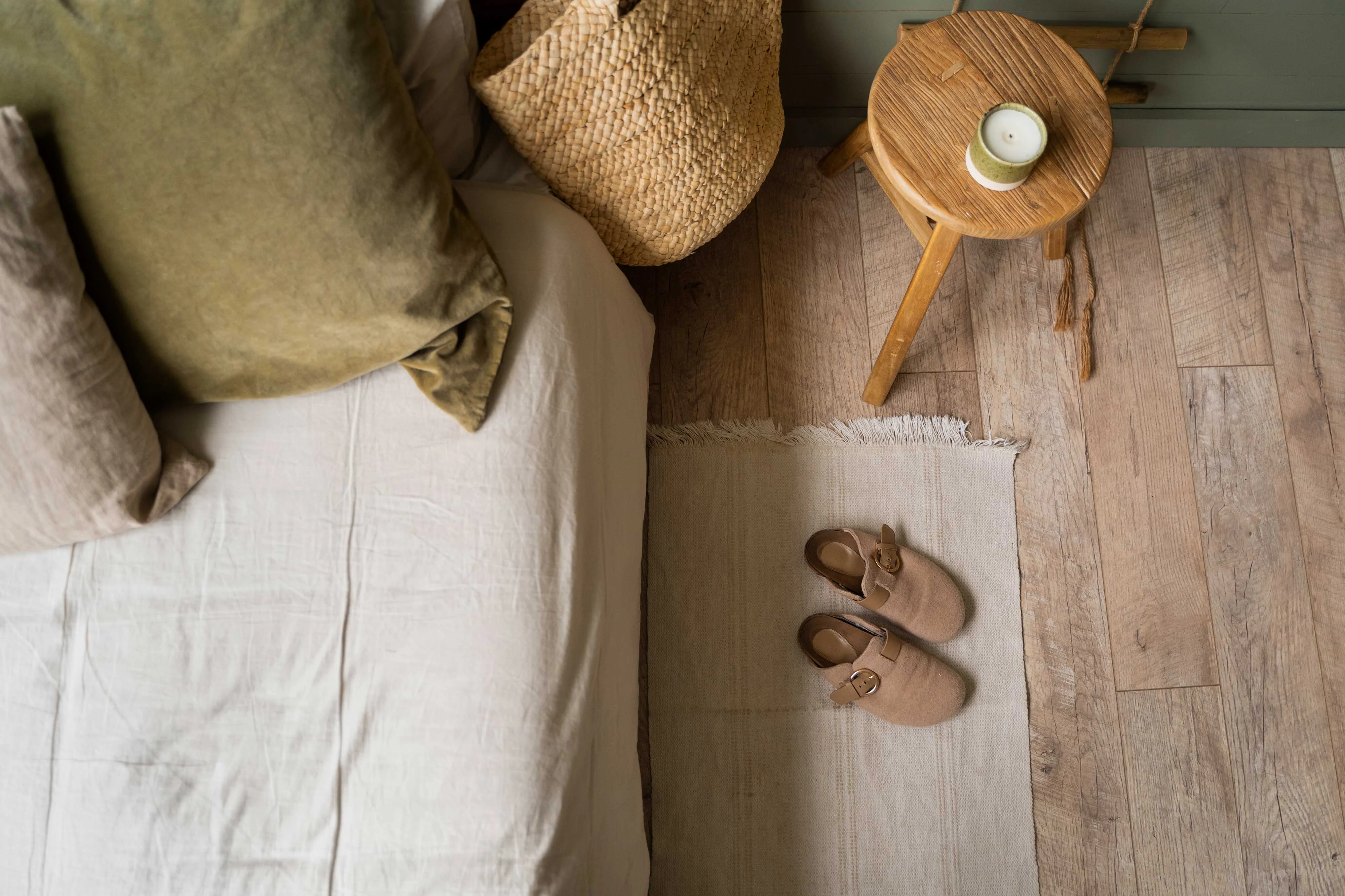 A cozy bedroom corner featuring a bed with textured linen bedding and green pillows, a woven basket, a small wooden stool with a candle, and a pair of slippers on a small rug.