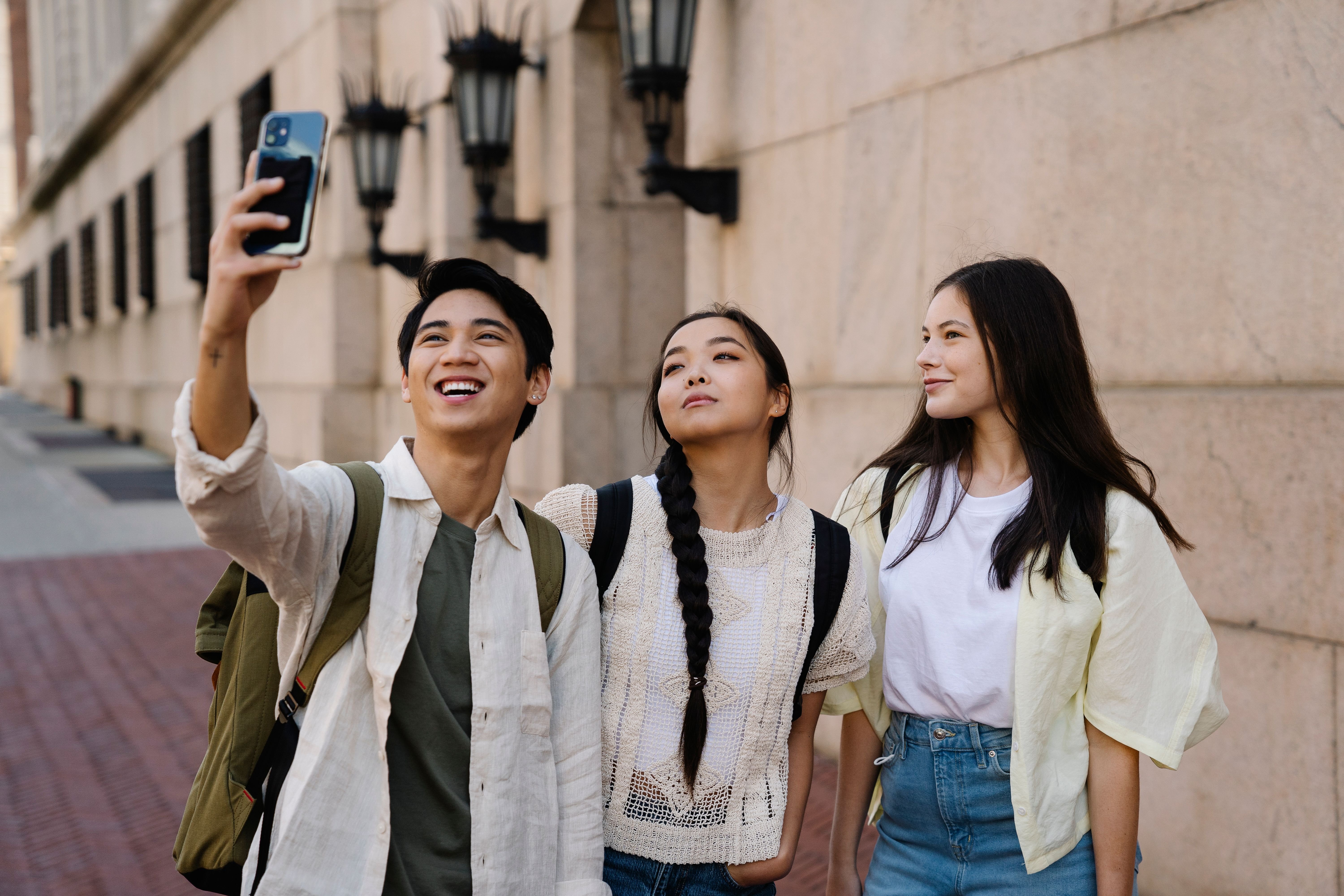 Three young adults taking a selfie together outdoors, with one person smiling and holding a smartphone, while the others strike a pose. They are in an urban setting with a stone wall and lanterns in the background.