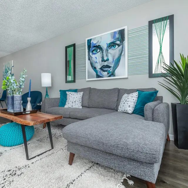 A stylish modern living room featuring a gray sectional sofa, a wooden coffee table, and decorative plants. The wall art includes a blue-toned face portrait, enhancing the contemporary aesthetic. The color scheme includes teal and gray, with a plush area rug on the floor.