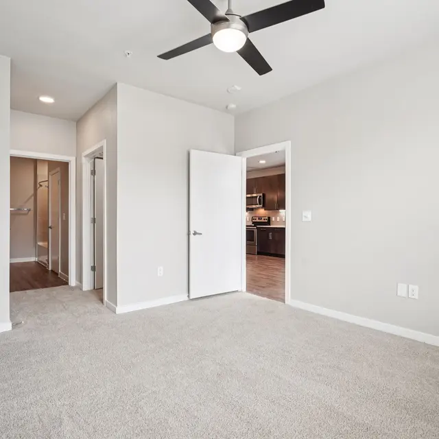 Spacious, modern apartment interior with beige carpet, ceiling fan, and open layout leading to kitchen area.