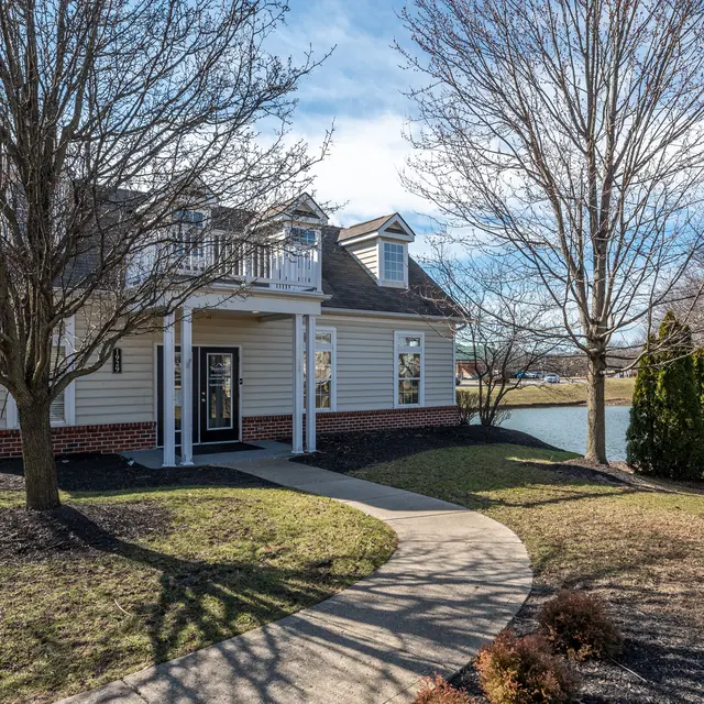 A charming home with a landscaped yard, featuring a winding path leading to the entrance. The house has a light-colored exterior with a balcony on the upper level and a scenic view of a pond. Leafless trees surround the property, indicating early spring or late fall.