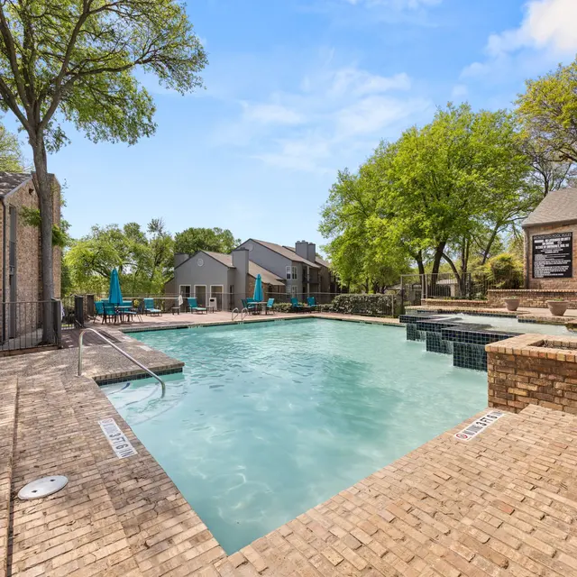 A well-maintained outdoor swimming pool area at an apartment complex, surrounded by trees and residential buildings. The pool is clear and inviting, with lounge chairs and umbrellas nearby.