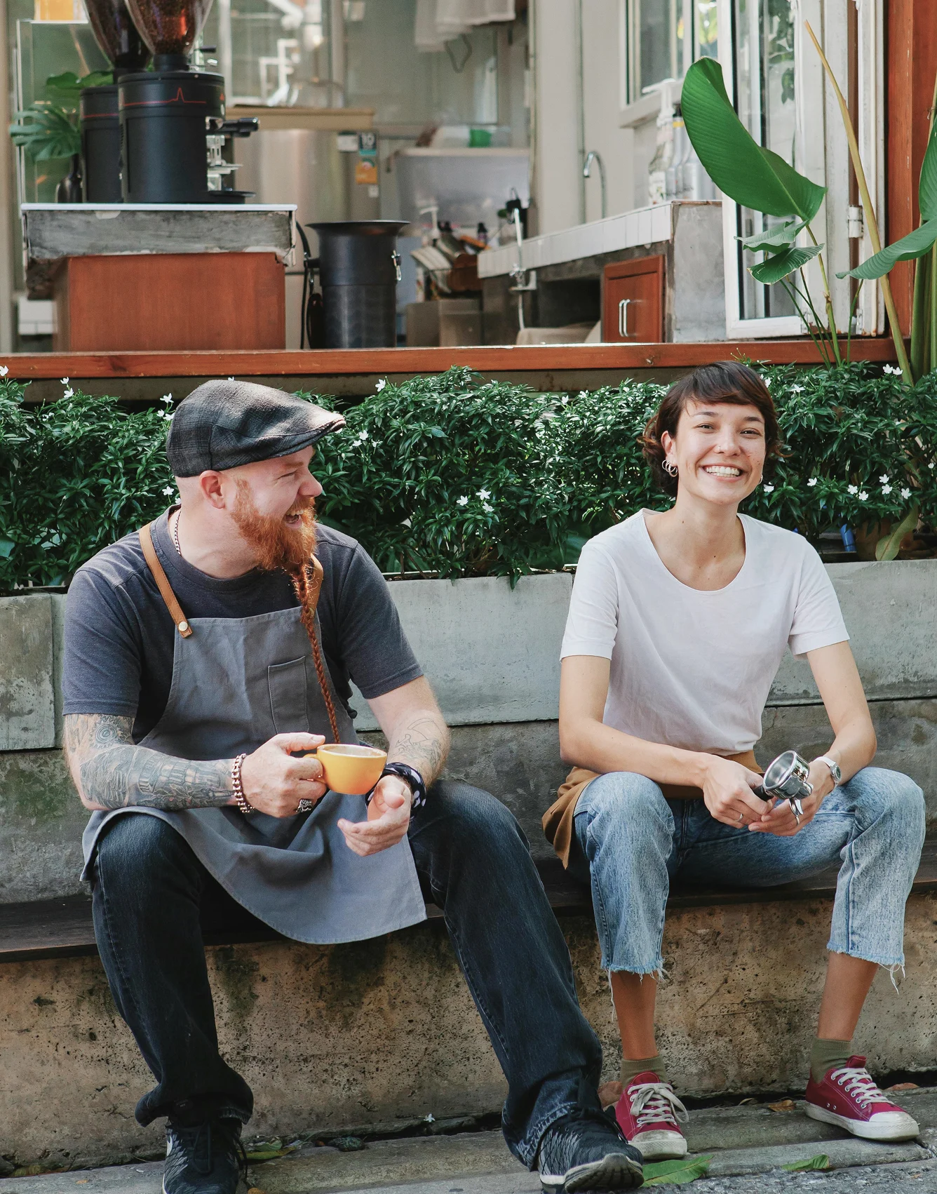 Coffee Shop Friends Two people sitting together outside a coffee shop, one holding a cup and the other holding a coffee grinder, both smiling.