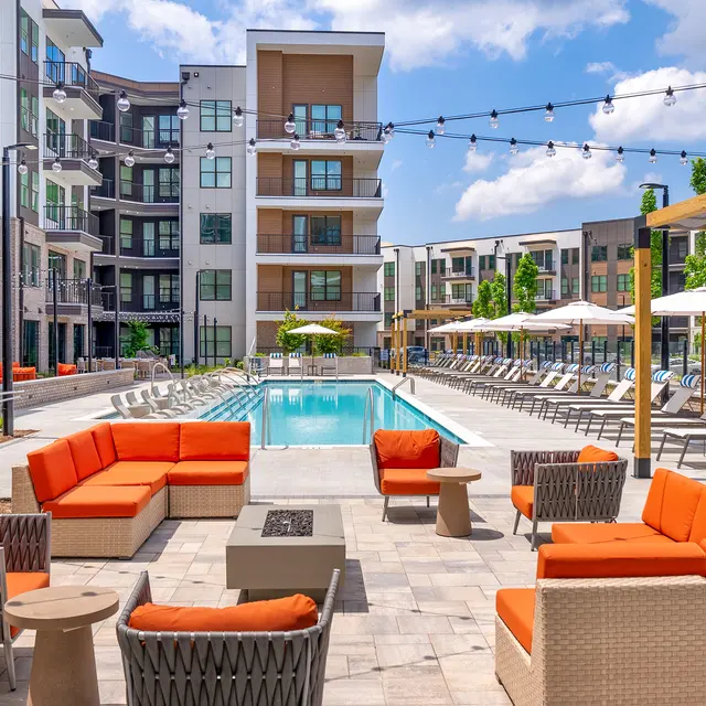 A modern apartment complex with a pool area. The patio features comfortable seating with orange cushions, a fire pit, and lounge chairs around a sparkling pool. Buildings are visible in the background under a partly cloudy sky.