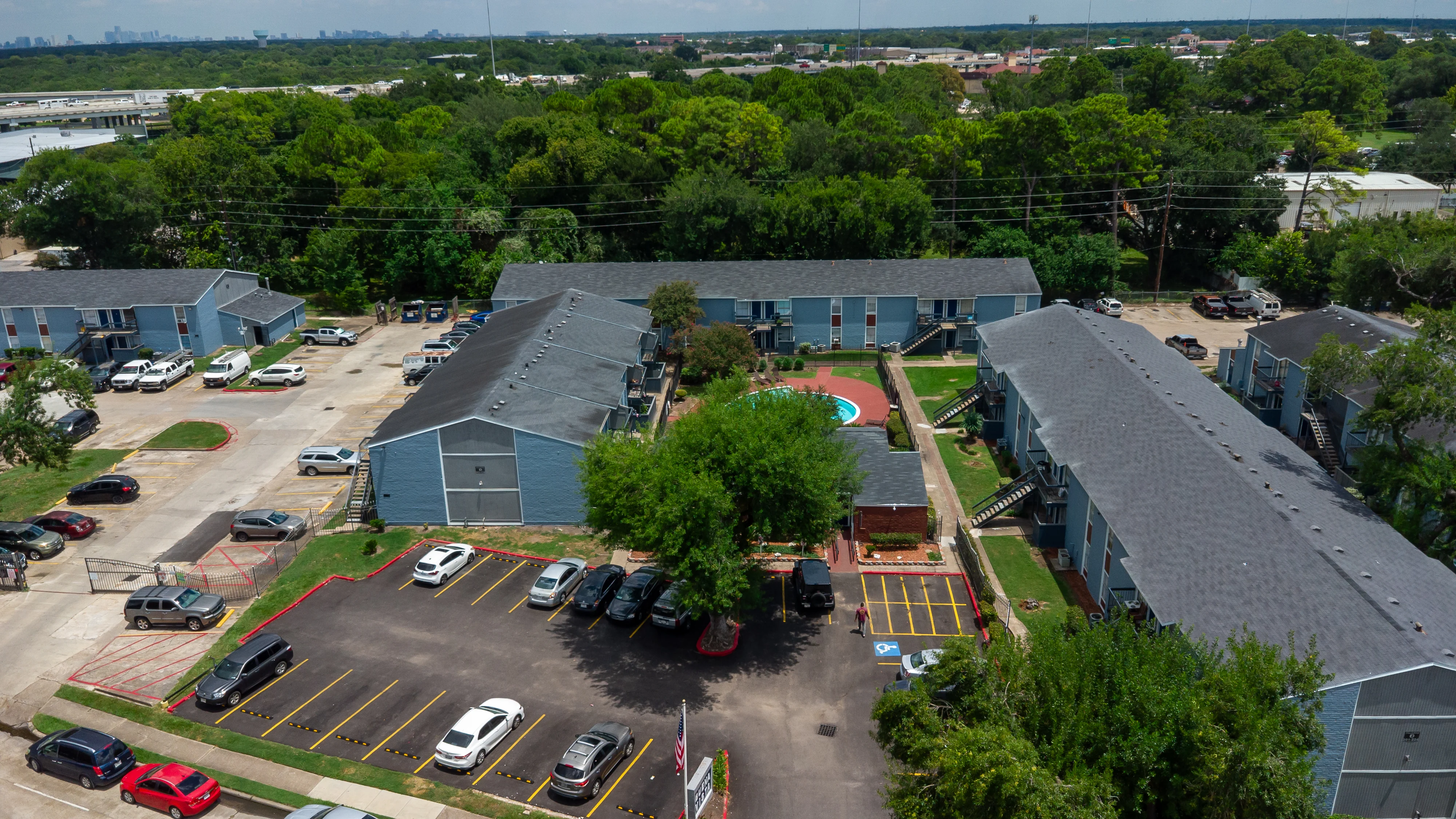 Aerial view of an apartment complex surrounded by greenery, featuring multiple buildings, parking spaces, and a playground area.