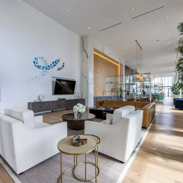 A contemporary living room featuring white armchairs, a round coffee table, a large television on the wall, and decorative plants. A wall mural adds a touch of color to the space.