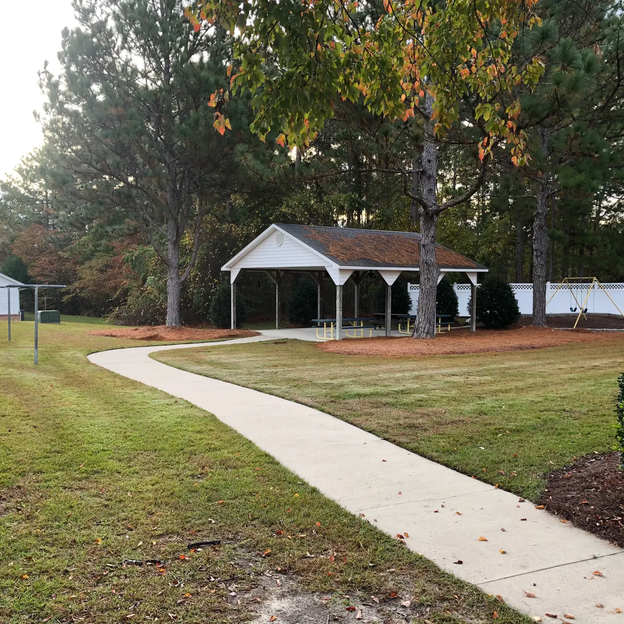 A landscaped backyard featuring a paved walkway, a white gazebo surrounded by green grass and trees, and a few outbuildings in the background.
