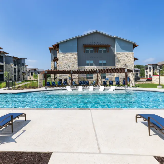 A spacious swimming pool surrounded by lounge chairs and an apartment building in the background on a sunny day.