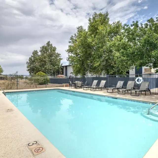 A clear swimming pool surrounded by lounge chairs and trees, under a partly cloudy sky.