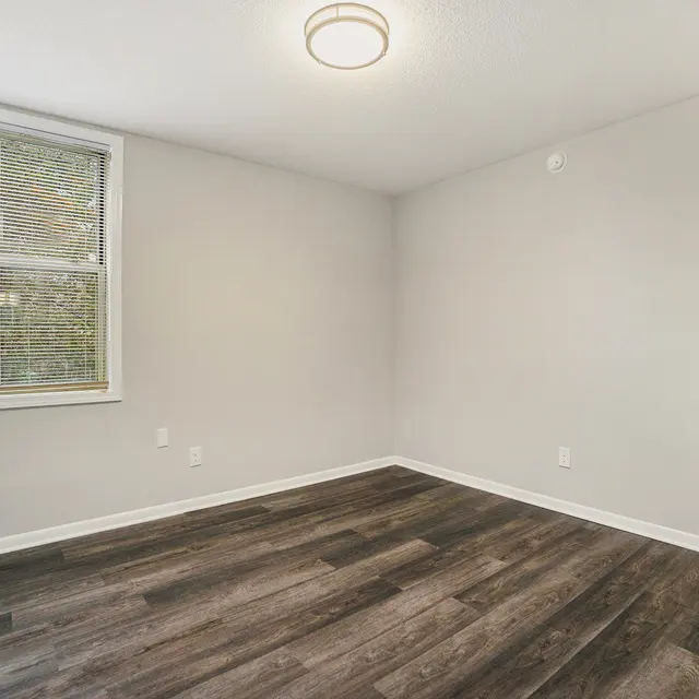 A vacant room with gray walls and dark wood flooring. A window with blinds is visible on one wall, and a ceiling light hangs in the center of the room.