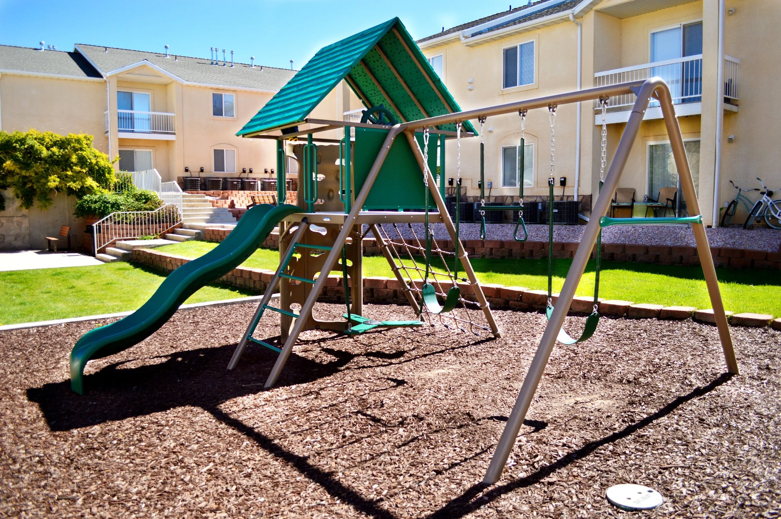Playground set with slides and swings in a residential area with apartment buildings in the background.