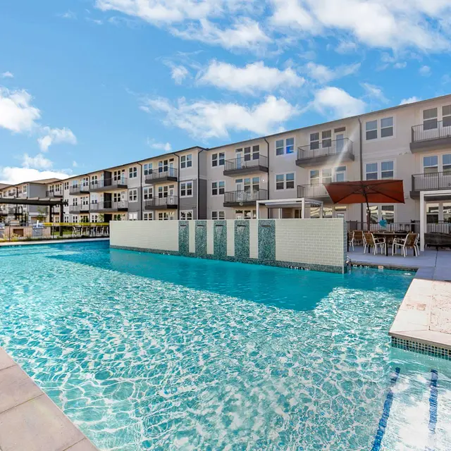 A modern apartment complex with a large swimming pool, surrounded by lounge areas and blue skies.