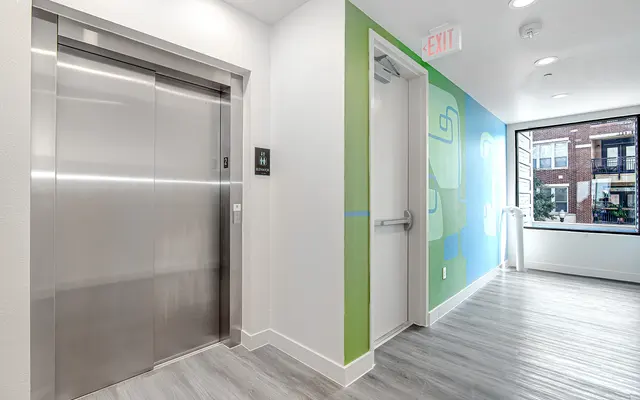 Interior view of a hallway featuring an elevator and a door. The wall to the right is painted green with abstract designs. A large window allows natural light to fill the space.