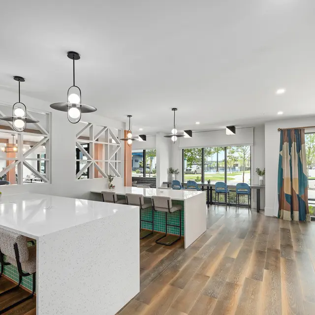 A modern interior of a spacious communal area with a white kitchen island, bar stools, large windows, and colorful decor.