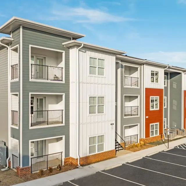 A modern multi-unit apartment building with a combination of beige, white, and red exterior. The building features balconies and multiple windows, with a clear blue sky in the background. Parking spaces are visible in front of the structure.