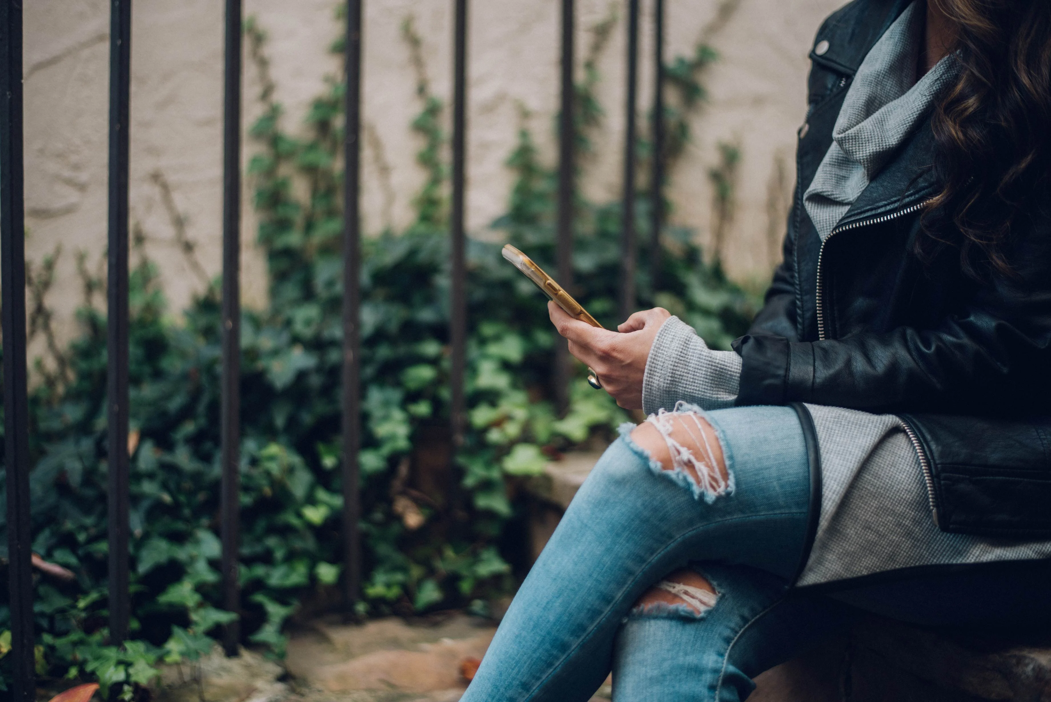 A person in a leather jacket and sweater sits on a bench, holding a phone, with greenery in the background. They're wearing ripped jeans.