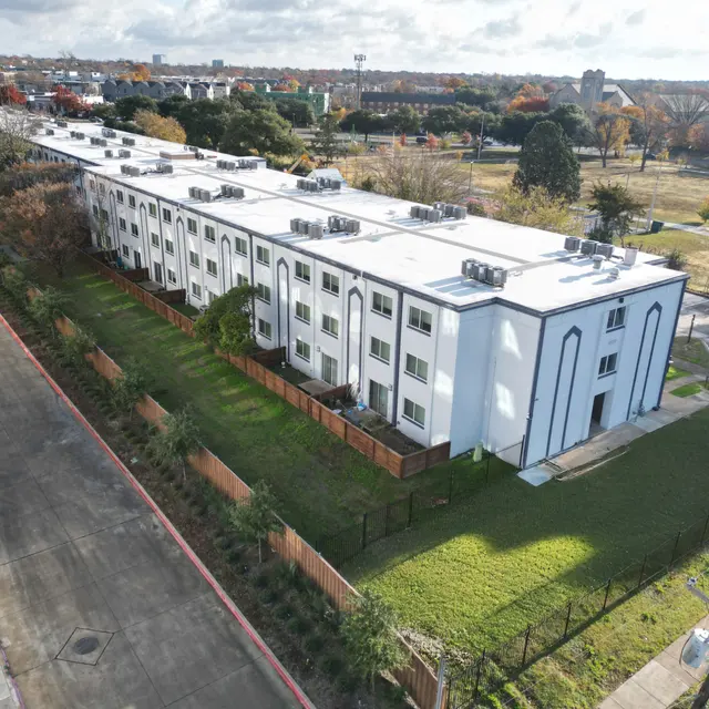 Aerial view of a modern apartment complex with multiple levels and a grassy area in front, surrounded by trees and a parking lot nearby.