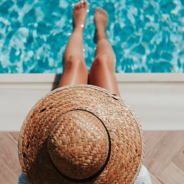A person wearing a straw hat sits by the edge of a pool, with their feet dangling above the shimmering water.