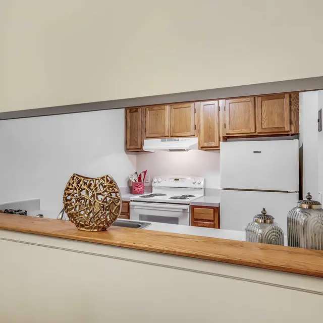 A view of a modern kitchen through an open wall pass. The kitchen features wooden cabinets, an electric stove, and white appliances. A decorative vase and two canisters are placed on the wooden ledge in front of the opening.