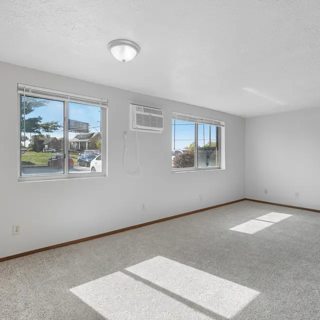 A bright and empty room with carpeted flooring, featuring two windows allowing natural light, and an air conditioning unit mounted on the wall.
