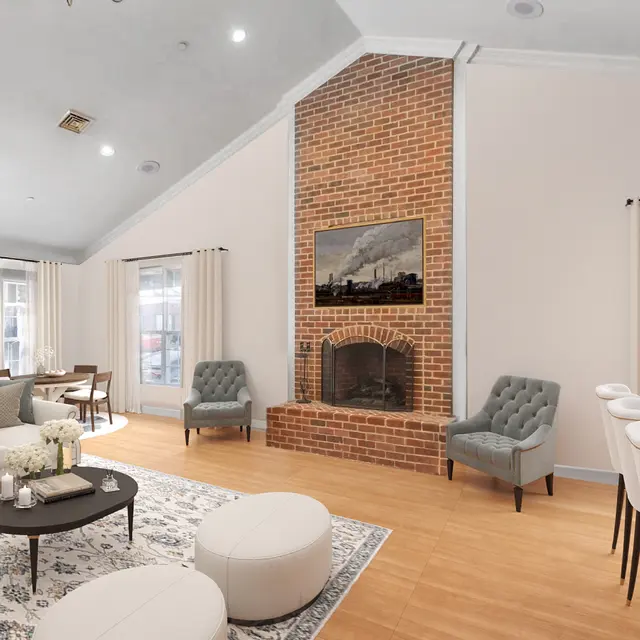 A modern living room featuring a large light-colored sofa, a round table with decorative flowers, and a brick fireplace. The space includes an elegant dining area with white chairs and a side table, complemented by large windows and light-colored curtains.