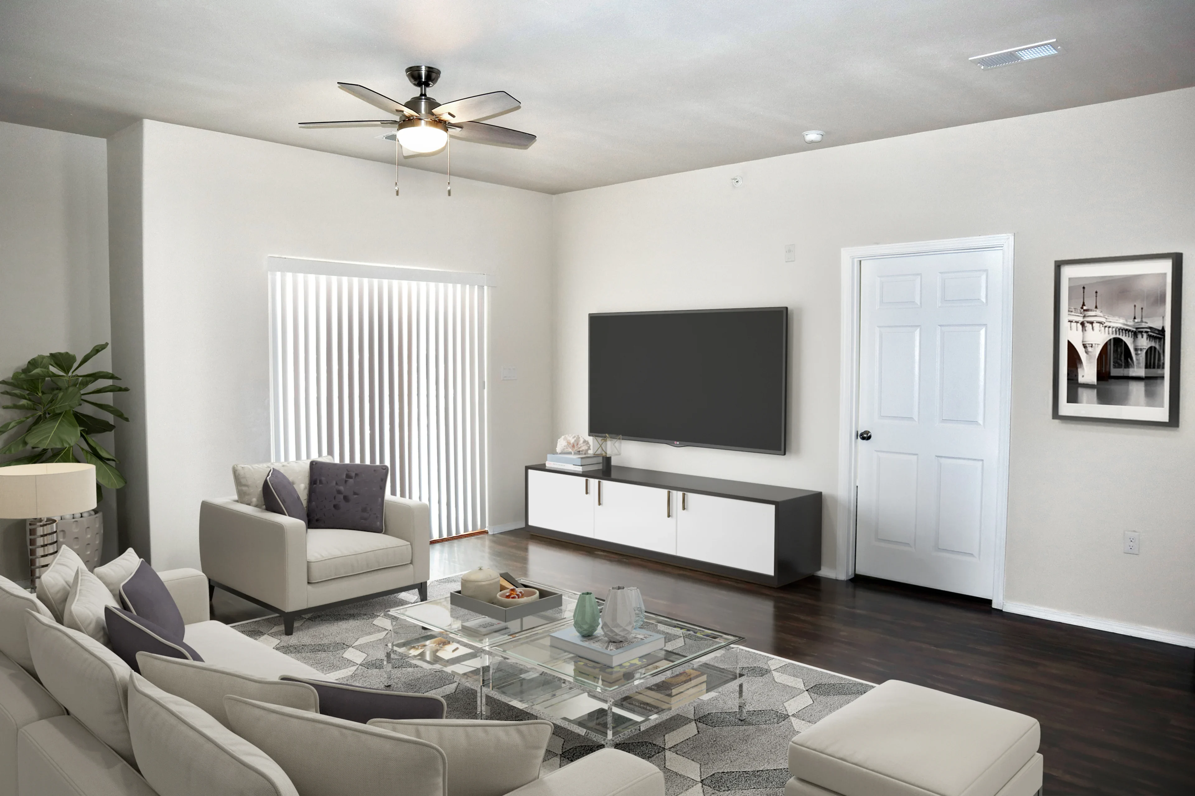 A modern living room featuring a light-colored sofa set with decorative pillows, a glass coffee table, and a television mounted on the wall across from the seating area. Natural light filters through vertical blinds on a window. A potted plant is visible in the corner. The walls are neutral-colored, and there is a door and a framed picture on the wall.