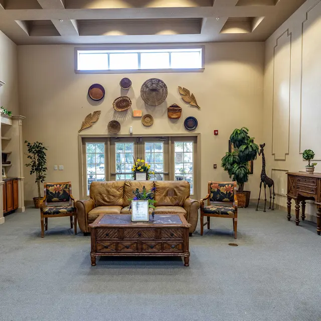 A spacious lobby area with a cozy seating arrangement, featuring a large brown sofa and decorative chairs. The wall displays various decorative items, and large windows allow natural light to flood in. There are potted plants and wooden cabinets enhancing the warmth of the space.